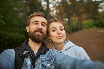 Fototapeta premium A caucasian couple in love takes a selfie in the park, against the backdrop of trees. A guy and a girl take a selfie close-up. A guy in a denim jacket and a girl in a hoodie take a selfie