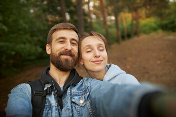 A caucasian couple in love takes a selfie in the park, against the backdrop of trees. A guy and a girl take a selfie close-up. A guy in a denim jacket and a girl in a hoodie take a selfie