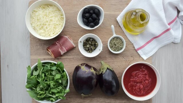Ingredientes de receta. Aceite, aceitunas negras, queso rallado, jamón serrano, alcaparras, orégano, rúcula, berenjenas y tomate triturado.