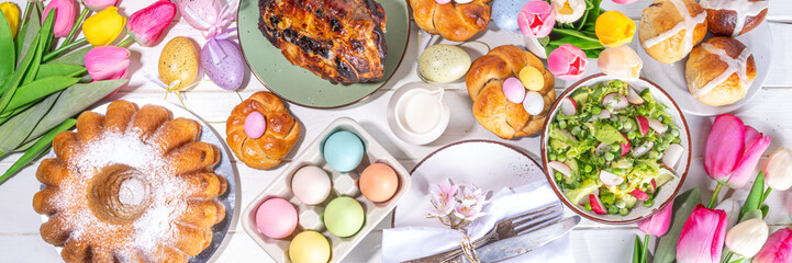Festive served Easter dinner or brunch table, with Easter egg, tulip flower bouquet, traditional Easter foods - cake, cross buns, wreath bread, glazed ham and spring salad, white table background