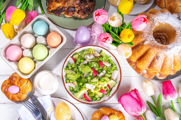 Festive served Easter dinner or brunch table, with Easter egg, tulip flower bouquet, traditional Easter foods - cake, cross buns, wreath bread, glazed ham and spring salad, white table background