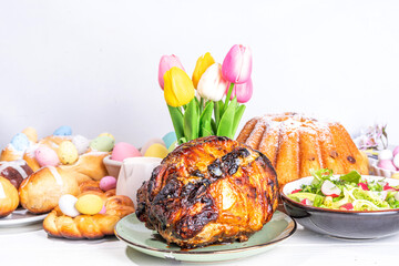 Festive served Easter dinner or brunch table, with Easter egg, tulip flower bouquet, traditional Easter foods - cake, cross buns, wreath bread, glazed ham and spring salad, white table background