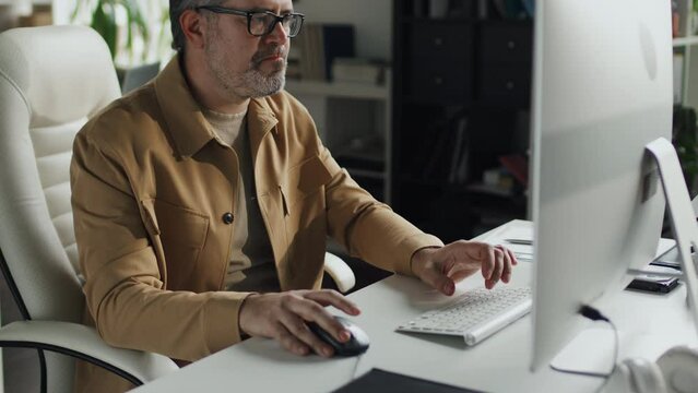 Tilt up shot of mature programmer in eyeglasses sitting at his workplace in front of computer monitor and typing codes on computer