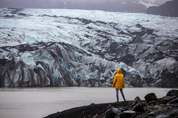 Girl in yellow raincoat standing on black sand near a lake, looking at the mighty blue outer...