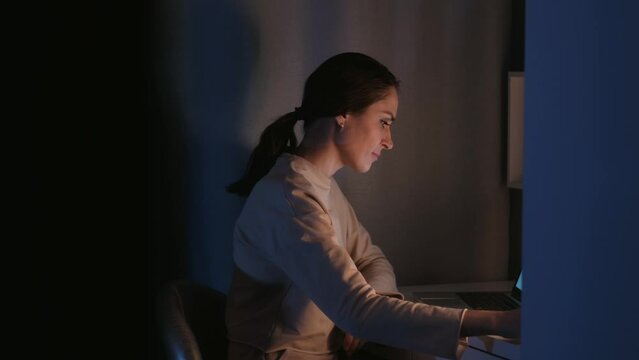 Young female looking in smartphone and use laptop at evening on table at home. Middle shot. 4k slowmotion.