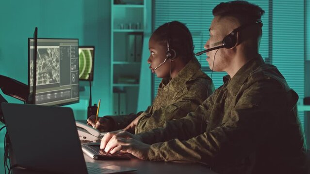 Two diverse male and female military surveillance officers in camouflage uniform spying target from satellite cameras on computer displays and controlling mission completion in dark office hub