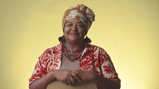 Portrait of cheerful mature African American woman in bright turban and Hawaiian print shirt posing for camera on yellow background