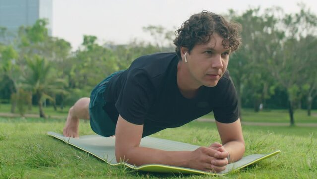 Man in sportswear and wireless earphones doing plank exercise on fitness mat during outdoor workout in park