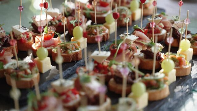 Neat bruschettas on French baguette with prosciutto, cheese and herbs are laid out on table and decorated with skewers with beads and hearts. Sunbeam falls through foliage on sandwiches.