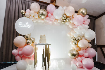 Arch decorated with pink, white and golden balloons, text baby. Trendy cake with figure angel. Copy space. Celebration baptism concept. Reception at birthday party on photo wall. Delicious reception.