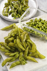 Peeling fresh steamed green soy beans edamame. Japanese plant snack on marble board on light grey background