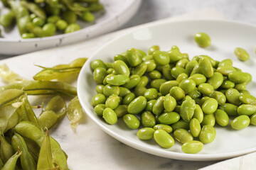 Peeling fresh steamed green soy beans edamame. Japanese plant snack on marble board on light grey background