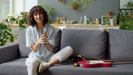 Gen Z girl blogger sitting on sofa at home, talking on camera and playing music with shaker while giving online class or filming vlog