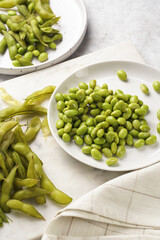 Peeling fresh steamed green soy beans edamame. Japanese plant snack on marble board on light grey background