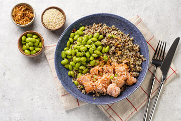 A blue bowl with quinoa tricolore, roughly chopped and pulled salmon and steamed green soy beans edamame with sesame seeds and deep fried onions on light background. Top view
