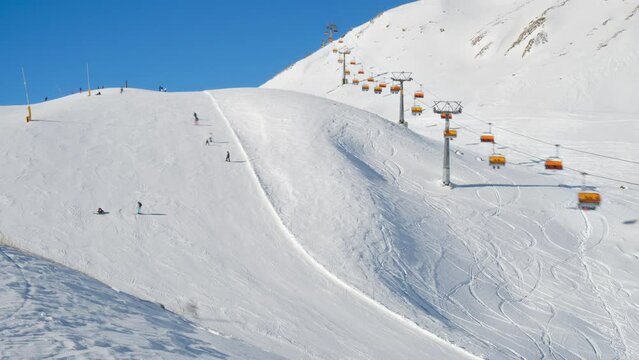 Timelapse of skiers on piste next to orange chair lift in Samnaun - Ischgl - Paznaun ski resort, located in Austria and Switzerland. Activity, sport, snow, winter, tourists.