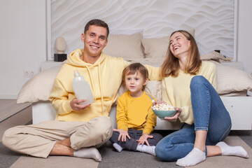 Cute adorable smiling parents with child son sitting by the bed and eating popcorn