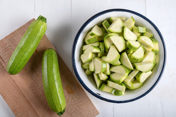 Two green zucchini on a cutting board with chopped zucchini in a bowl on a wooden background.