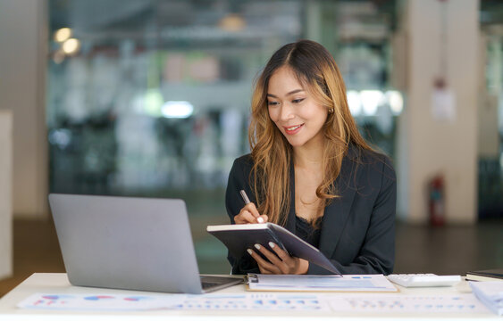 Beautiful Asian businesswoman using her laptop to work and enjoy taking notes on meeting minutes agendas of secretarial work.