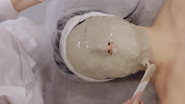 Cosmetologist doctor making medical peeling face mask in spa salon. Beautician applying mud cream on middle-aged woman face. Girl skin care procedure. Facial cosmetology treatment. Vertical view shot