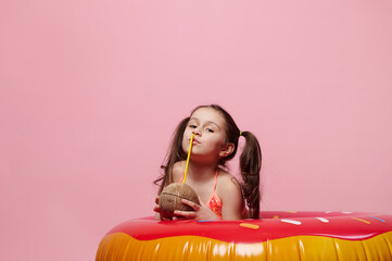 Cute little girl in swimsuit drinking coconut cocktail, posing with donut inflatable ring, isolated pink background