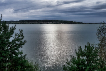 Pine trees grow on the banks of a large river, a beautiful cloudy sky, the surface of the water reflects sunlight with light ripples, a beautiful summer landscape