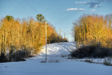 A clear-cut through the woods has utility lines running down the middle of it.  Sun is setting on the power lines and poles in this snow-covered field in Upstate NY this winter. 