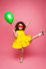 Happy birthday celebration with flying balloons of a charming cute little girl in a yellow dress isolated on a pink background.