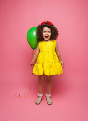 Happy birthday celebration with flying balloons of a charming cute little girl in a yellow dress isolated on a pink background.