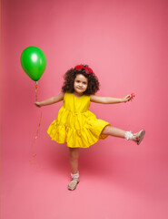 Happy birthday celebration with flying balloons of a charming cute little girl in a yellow dress isolated on a pink background.
