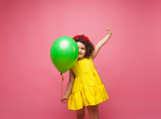 Happy birthday celebration with flying balloons of a charming cute little girl in a yellow dress isolated on a pink background.