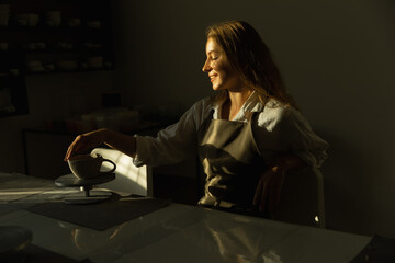a girl potter sits at a table, admiring her product, warm light.