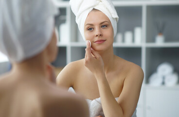 Young woman sits in the bathroom in front of the makeup mirror and does cosmetic procedures. Beautiful girl in white towel. Skin care, health, rejuvenation and spa treatment concept.