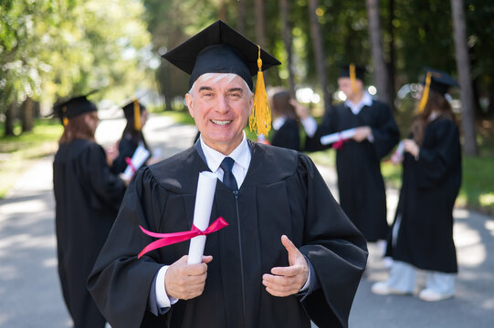 A Group Of Graduates In Robes Outdoors. An Elderly Student Rejoices At Receiving A Diploma.