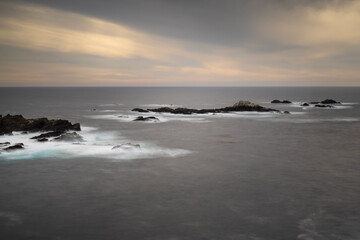 Fototapeta premium Sunset at Point Lobos State Natural Reserve, long exposure, featuring the ocean and rocks against a partly cloudy sky with orange hues. Carmel, California, USA.