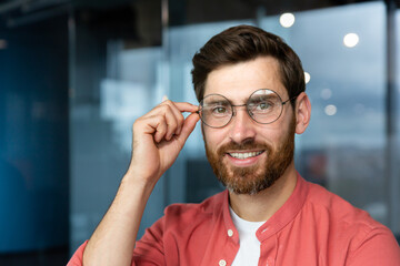 Close-up portrait of young businessman in red shirt, mature man in glasses smiling and looking at camera inside office near window, successful investor happy with achievements and good investment.