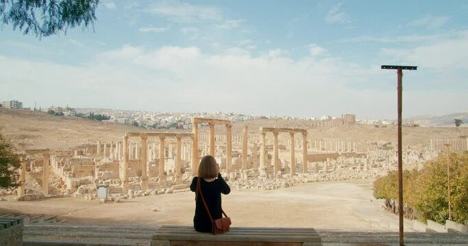 Young lady snapping pictures by analogue camera of ancient ruins landscape. Touristic woman sitting and taking photos of North Colonnaded Street in Gerasa, Jerash, Jordan. 4k zoom in shot