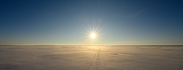 A cold looking winter scene with a large snow covered lake and a clear blue sky. A snowmobile track crosses the lake heading towards a bright sun.
