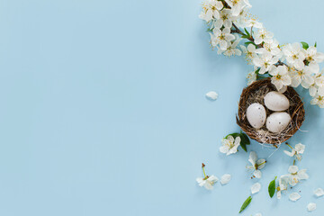Stylish Easter eggs in nest and cherry blossoms flat lay on blue background with copy space. Happy Easter! Greeting card template. Modern holiday banner. Festive minimal composition