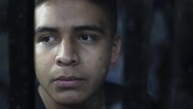 detailed view of the face of a young latin man, very pensive looking through a window. man behind bars, in a dark room.