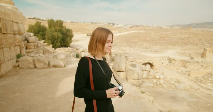 Touristic woman sightsees and takes photos of landscape and landmarks in Gerasa, Jerash, Jordan. Young lady snaps pictures by analogue camera among ancient ruins in summer. 4k trucking shot