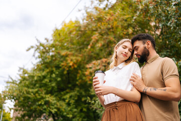 Low-angle view of happy young couple in love standing hugging with closed eyes at city park on background of trees. Tattooed handsome man and pretty female walking spending time together on summer day