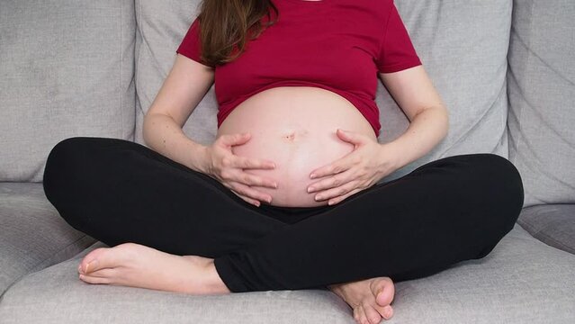 Unrecognizable pregnant woman stroking tummy sitting on sofa at home