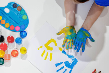 Children's handprints. Blue and yellow palm prints on a white sheet of paper, children's hands in paint, a palette of colorful paints, gouache. Top view of the table. Children's creativity