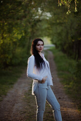 Sexy sensual brunette walks in a summer forest at sunset in a white blouse and jeans