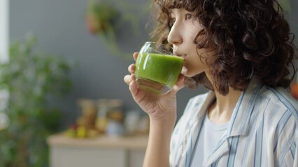 Tilt up shot of young woman drinking freshly made green smoothie from glass while standing in kitchen - Powered by Adobe