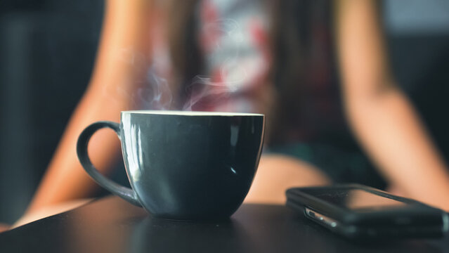 Close-up image of cup having fresh hot tea, coffee, drink laying on table with unrecognizable young girl in background sitting on sofa couch at home lifestyle. woman relaxing, enjoying morning hour