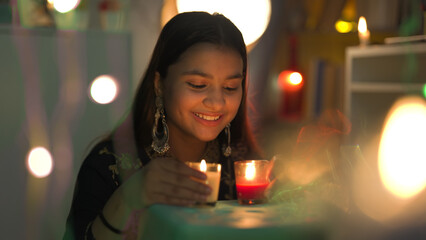 Smiling pretty girl wearing big earrings sitting and look burning glass candles closer to face on the occasion of Diwali. Indian teenage enjoy Deepavali festival with decorative surroundings at home