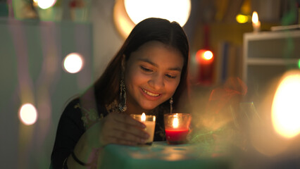 Smiling pretty female wearing big earrings sitting and hold burning glass candles in hand closer to face on the occasion of Diwali. Indian teenager enjoy Deepawali festival with Decorative surrounding