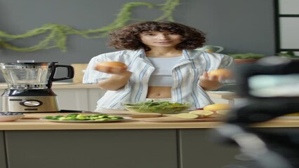 Vertical shot of vegan girl standing in kitchen and telling about fresh veggies while filming healthy food blog with digital camera